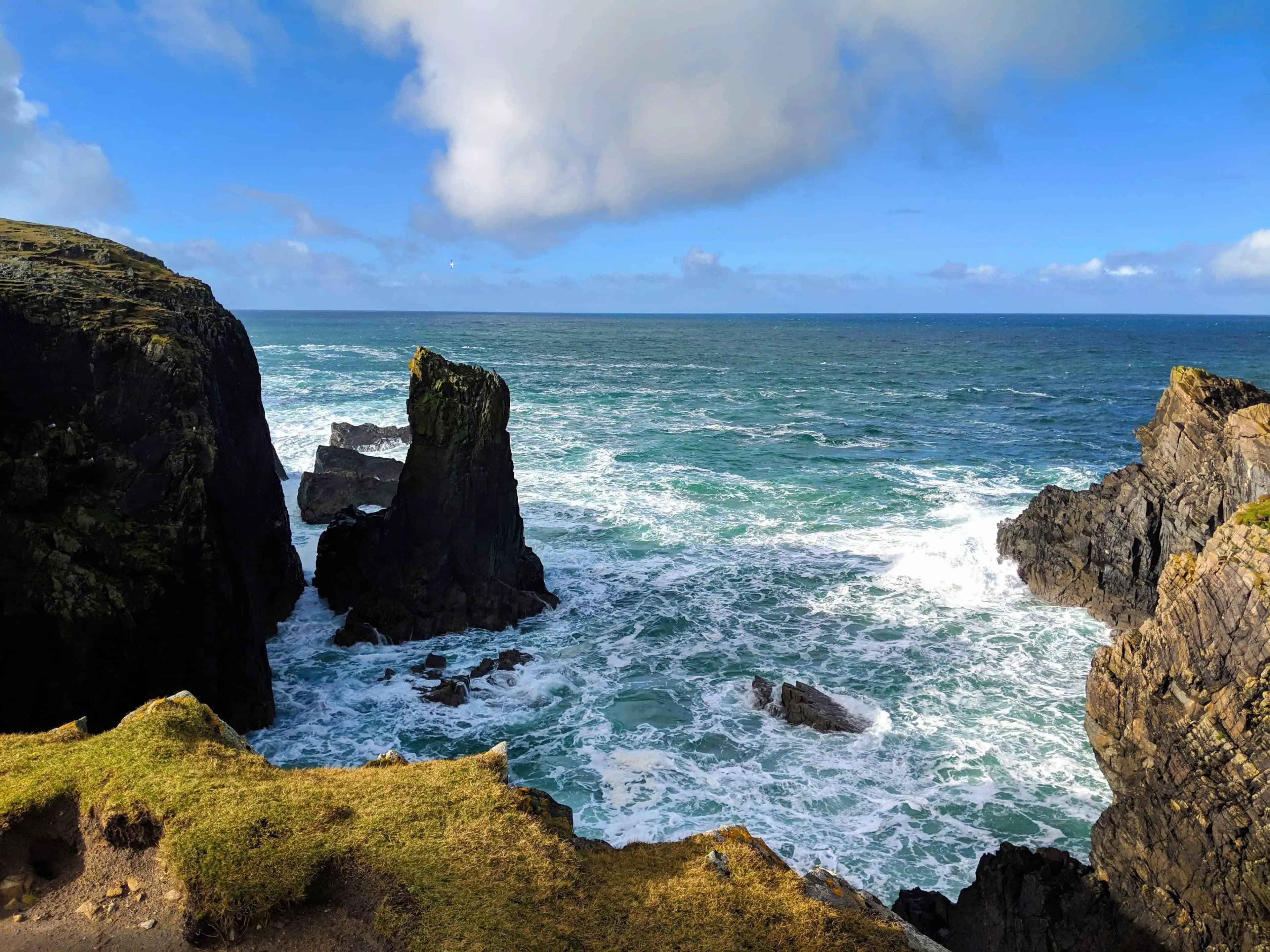 Butt of Lewis coastline with rugged cliffs, jagged sea stacks, and waves crashing into the Atlantic under a bright, partly cloudy sky.